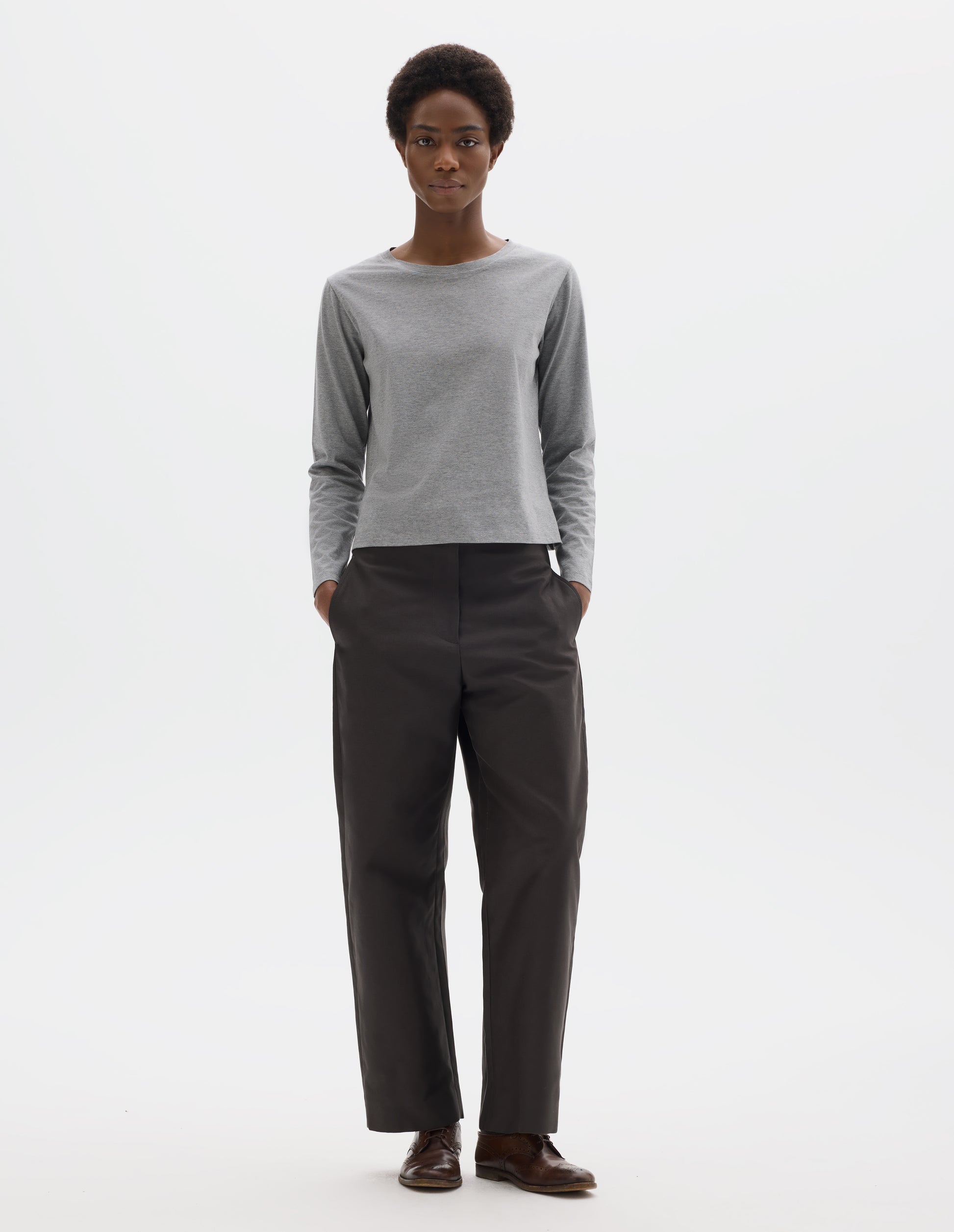Full body image, front view of model wearing grey relaxed cotton t-shirt with wide round neck, long sleeves and boxy cut. Styled with dark grey trousers and brown leather brogues.