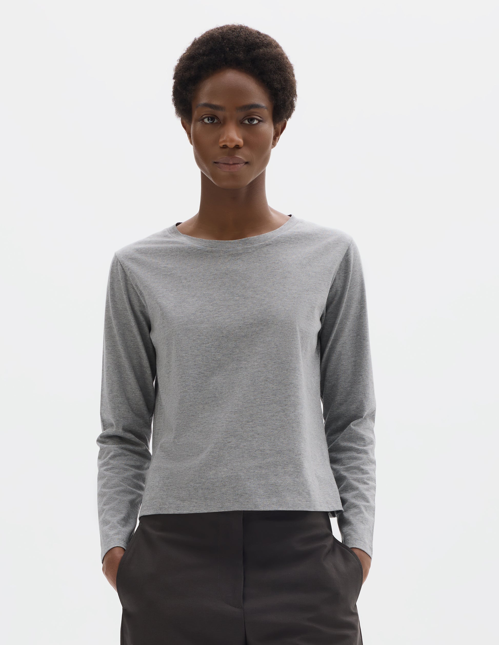 Cropped body image, front view of model wearing grey relaxed cotton t-shirt with wide round neck, long sleeves and boxy cut. Styled with dark grey trousers.