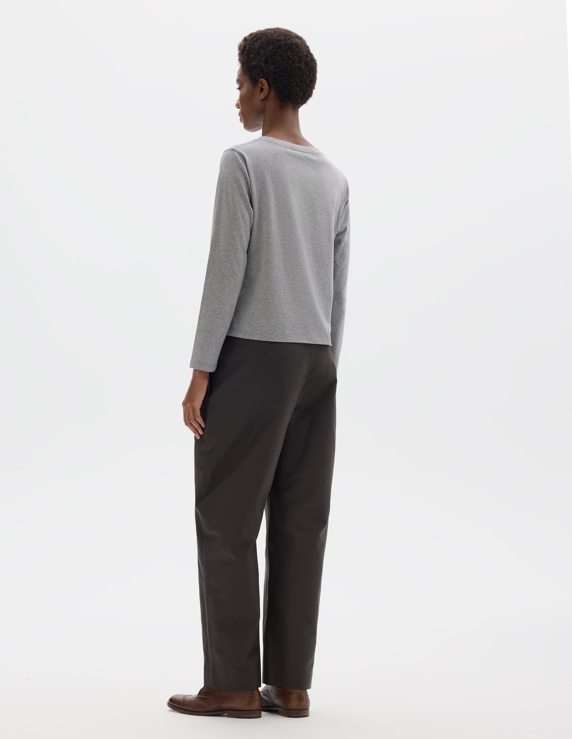 Full body image, back view of model wearing grey relaxed cotton t-shirt with wide round neck, long sleeves and boxy cut. Styled with dark grey trousers and brown leather brogues.