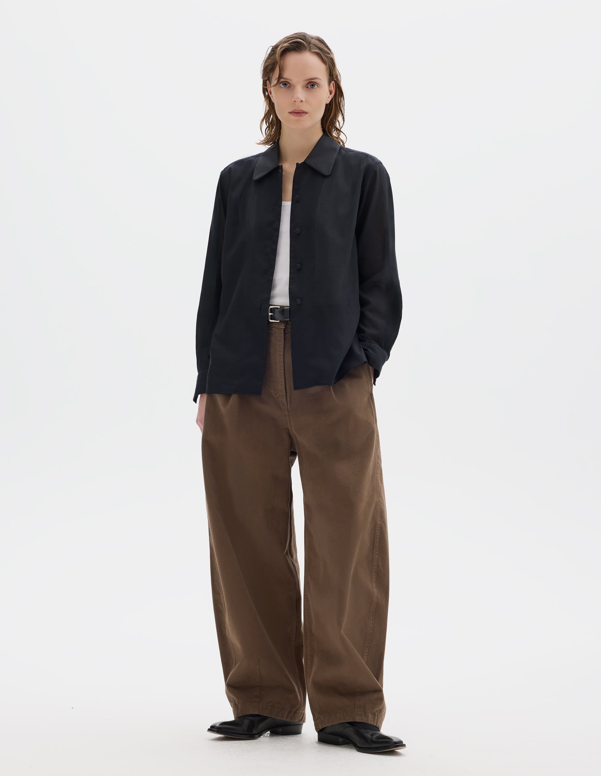 Full body image, front view of model wearing black cotton cashmere shirt with contrast cotton poplin collar, covered domed buttons on placket and cuffs, and with deep vent split hem. Styled over white vest, and with dark beige trousers with black leather belt and black leather shoes.
