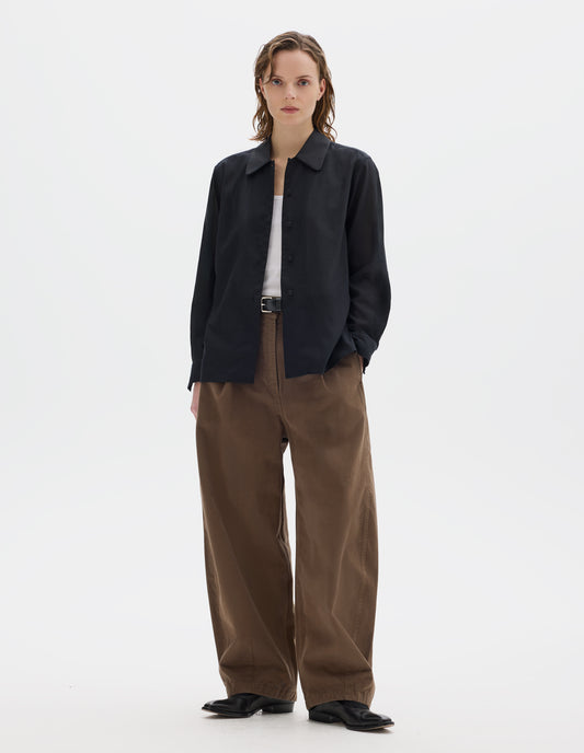 Full body image, front view of model wearing black cotton cashmere shirt with contrast cotton poplin collar, covered domed buttons on placket and cuffs, and with deep vent split hem. Styled over white vest, and with dark beige trousers with black leather belt and black leather shoes.