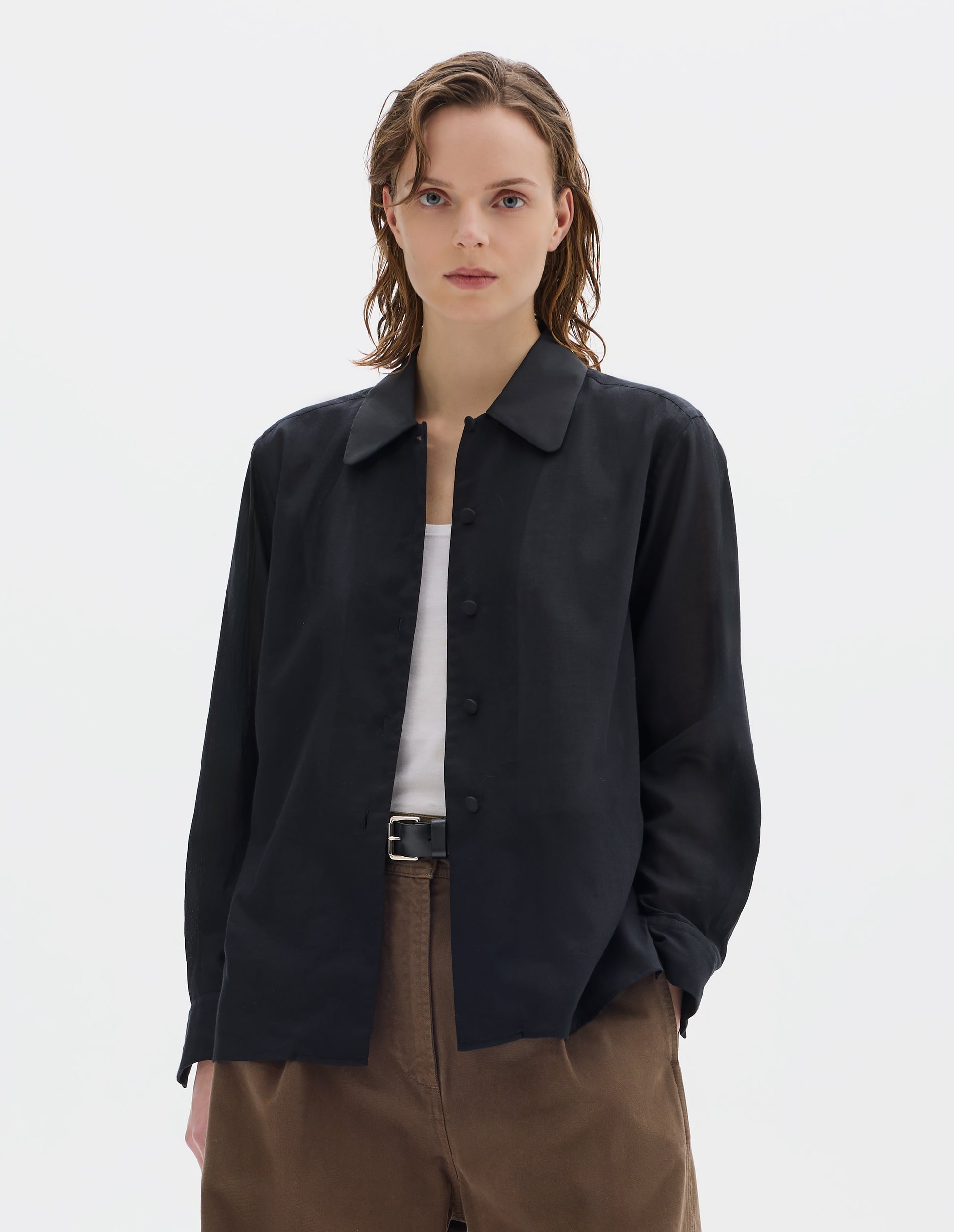 Cropped body image, front view of model wearing black cotton cashmere shirt with contrast cotton poplin collar, covered domed buttons on placket and cuffs, and with deep vent split hem. Styled over white vest, and with dark beige trousers with black leather belt.