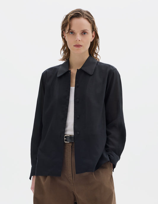 Cropped body image, front view of model wearing black cotton cashmere shirt with contrast cotton poplin collar, covered domed buttons on placket and cuffs, and with deep vent split hem. Styled over white vest, and with dark beige trousers with black leather belt.
