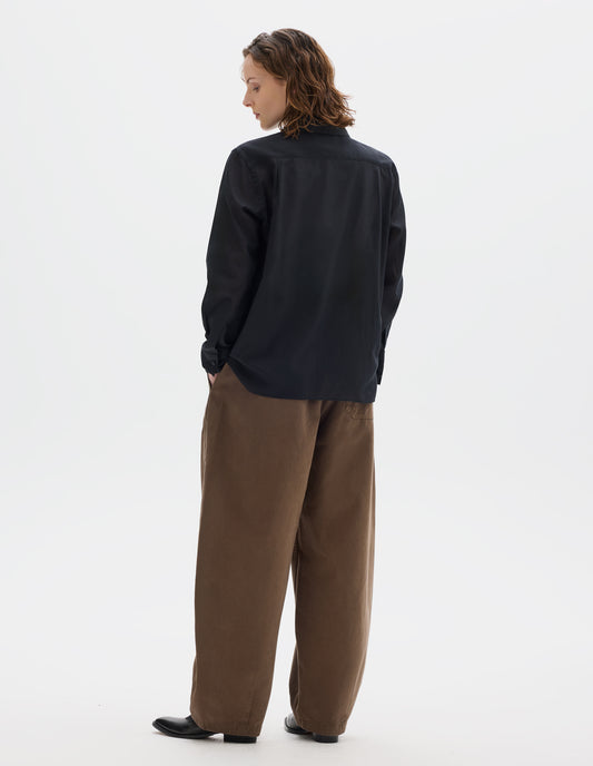 Full body image, back view of model wearing black cotton cashmere shirt with contrast cotton poplin collar, covered domed buttons on cuffs, and deep vent split hem. Styled with dark beige trousers and black leather shoes.
