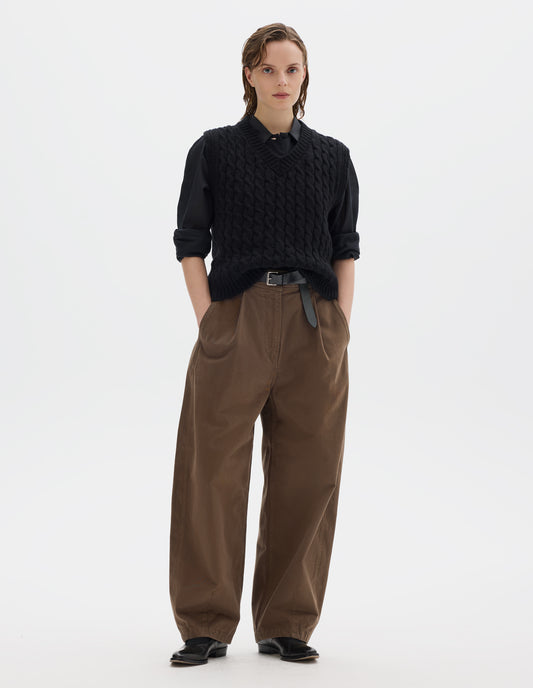 Full body image, front view of model wearing relaxed black wool slipover with V neck, front cable design and ribbed neck, armholes, side seams and hem. Styled over black black shirt, and with brown trousers with black leather belt and black leather shoes.
