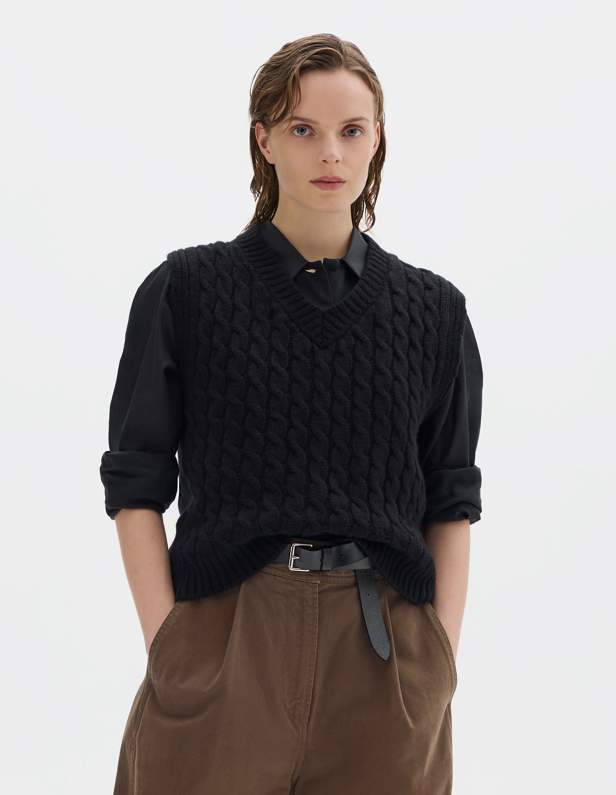 Cropped body image, front view of model wearing relaxed black wool slipover with V neck, front cable design and ribbed neck, armholes, side seams and hem. Styled over black black shirt, and with brown trousers with black leather belt.