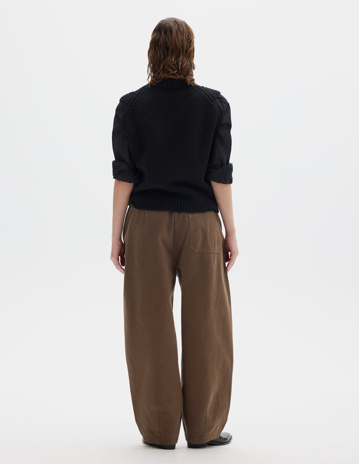 Full body image, back view of model wearing relaxed black wool slipover with plain knitted back and ribbed neck, armholes, side seams and hem. Styled over black black shirt, and with brown trousers and black leather shoes.
