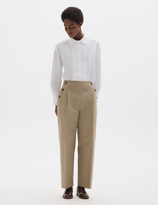  Full body image, front view of model wearing beige cotton linen cropped trousers with pleats, horn button side cinches and three horn button closures on both sides, and with stitch-through hem. Styled with white shirt and brown leather brogues.