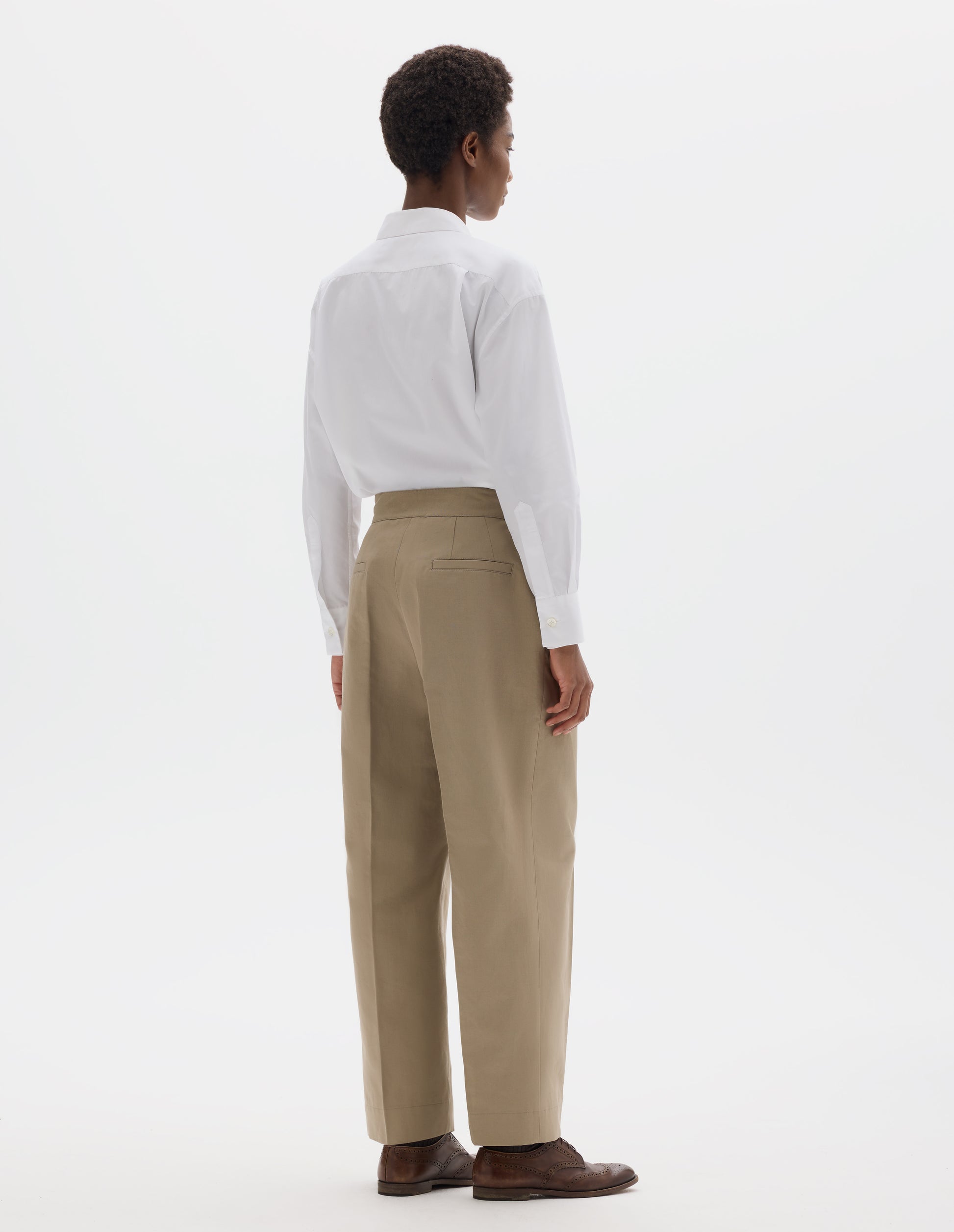 Full body image, back view of model wearing beige cotton linen cropped trousers with side cinches and welt back pockets. Styled with white shirt and brown leather brogues.