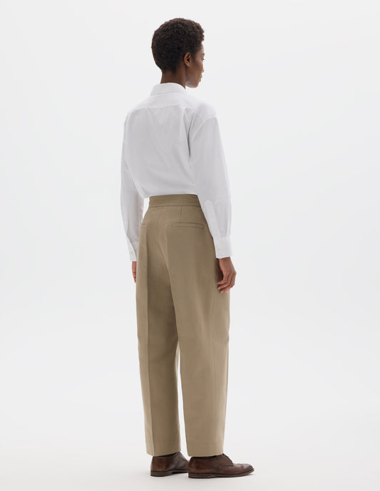 Full body image, back view of model wearing beige cotton linen cropped trousers with side cinches and welt back pockets. Styled with white shirt and brown leather brogues.
