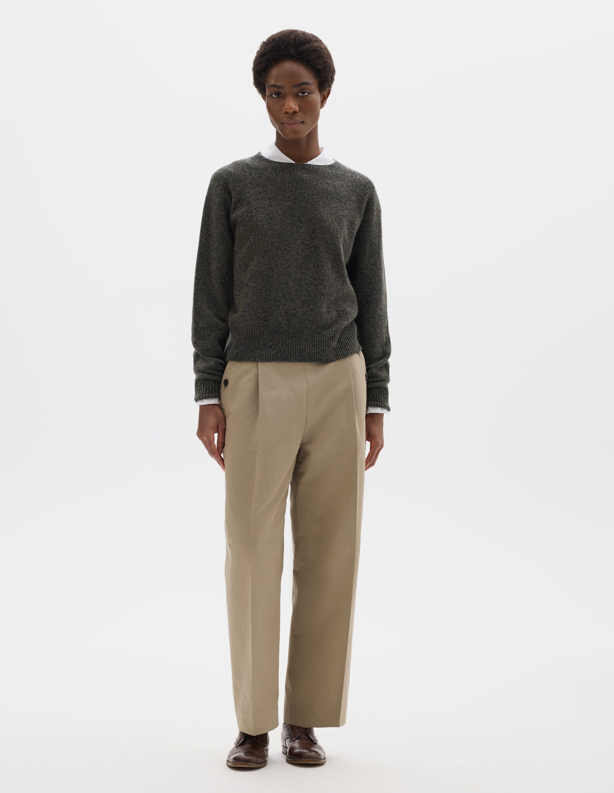 Full body image, front view of model wearing brownish grey fully fashioned seamless merino cashmere  jumper with crew neck, raglan sleeves, and with ribbed neck, cuffs and hem. Styled over white shirt, and with beige trousers and brown leather brogues.