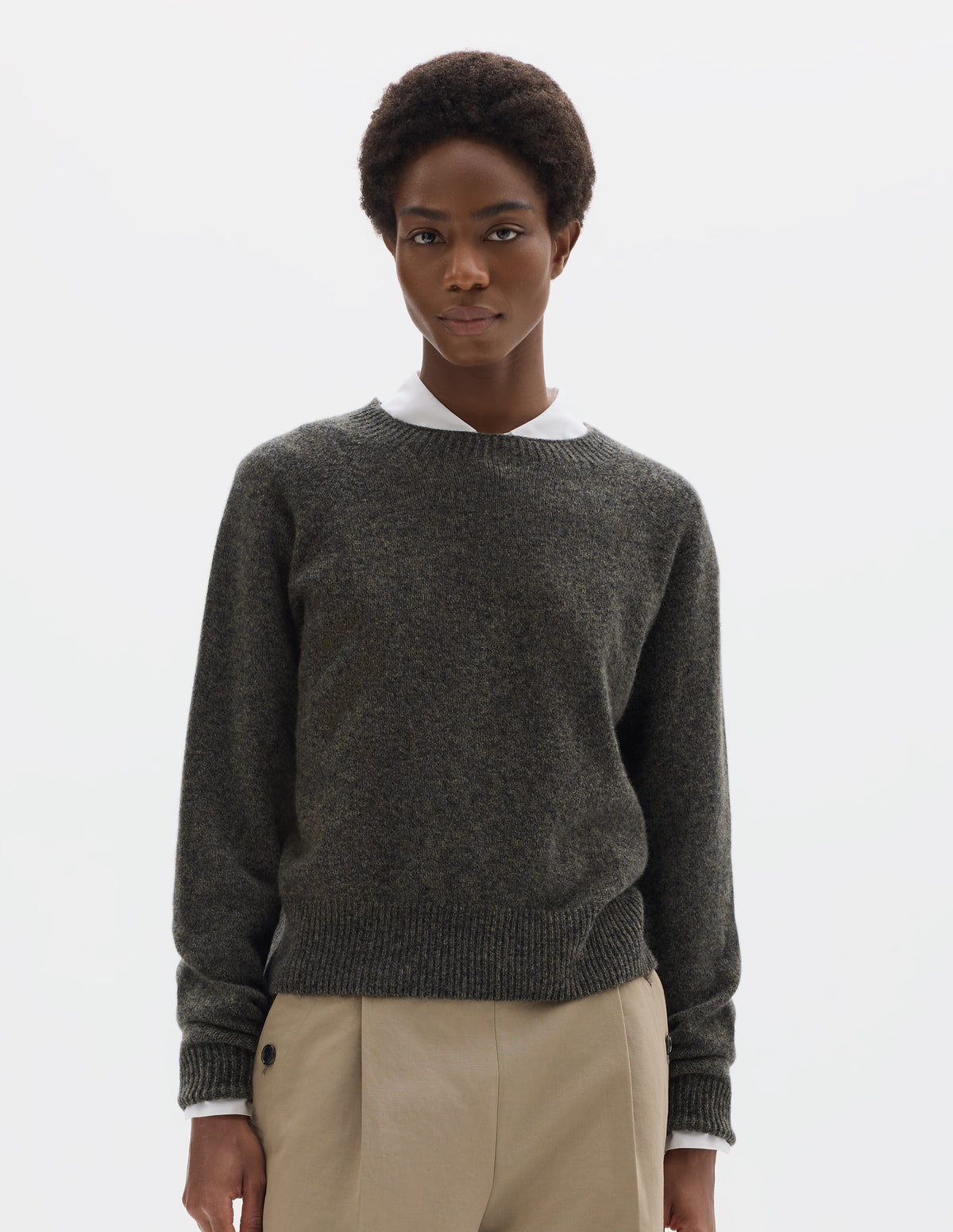 Cropped body image, front view of model wearing brownish grey fully fashioned seamless merino cashmere jumper with crew neck, raglan sleeves, and with ribbed neck, cuffs and hem. Styled over white shirt, and with beige trousers.