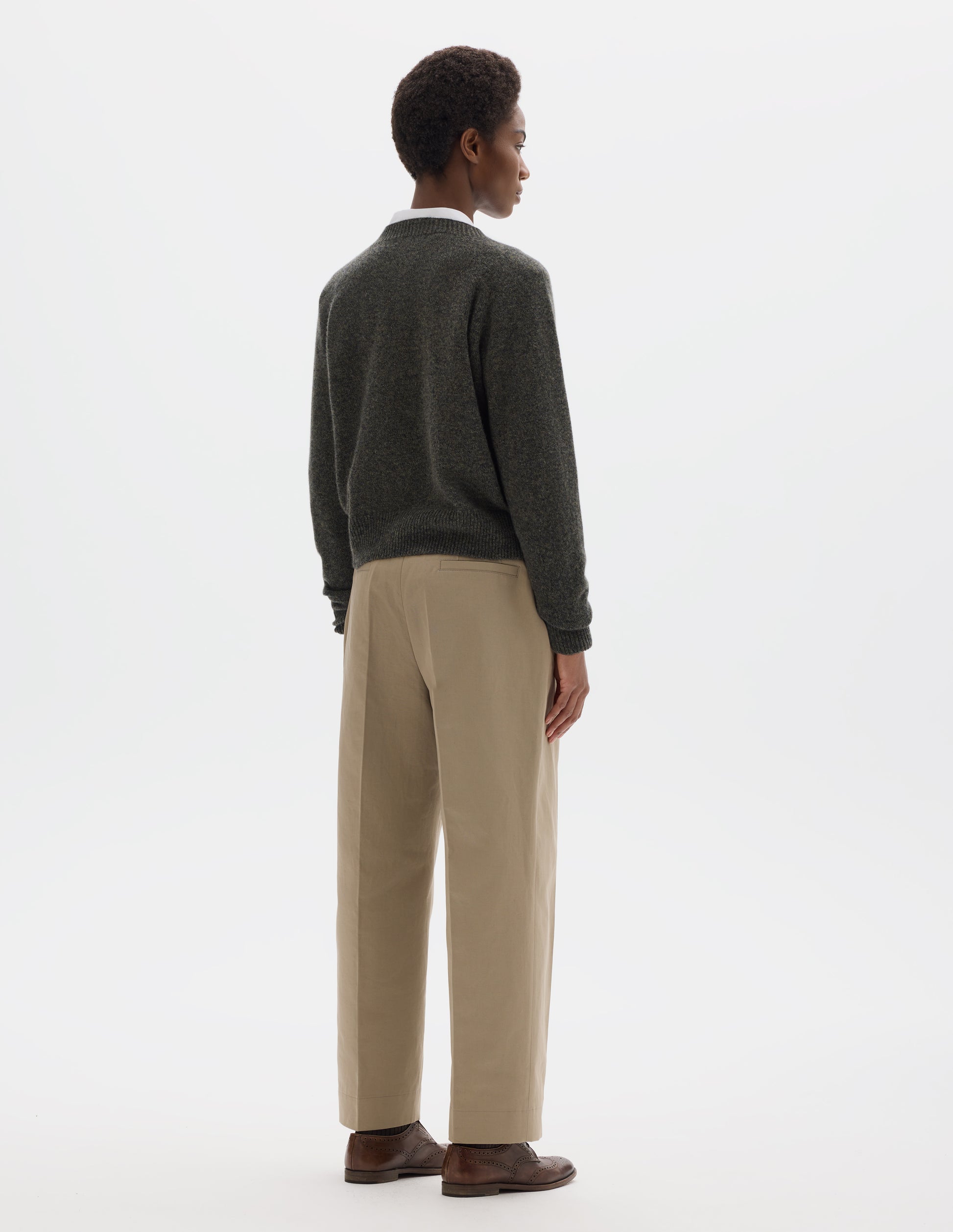 Full body image, back view of model wearing brownish grey fully fashioned seamless merino cashmere  jumper with crew neck, raglan sleeves, and with ribbed neck, cuffs and hem. Styled over white shirt, and with beige trousers and brown leather brogues.