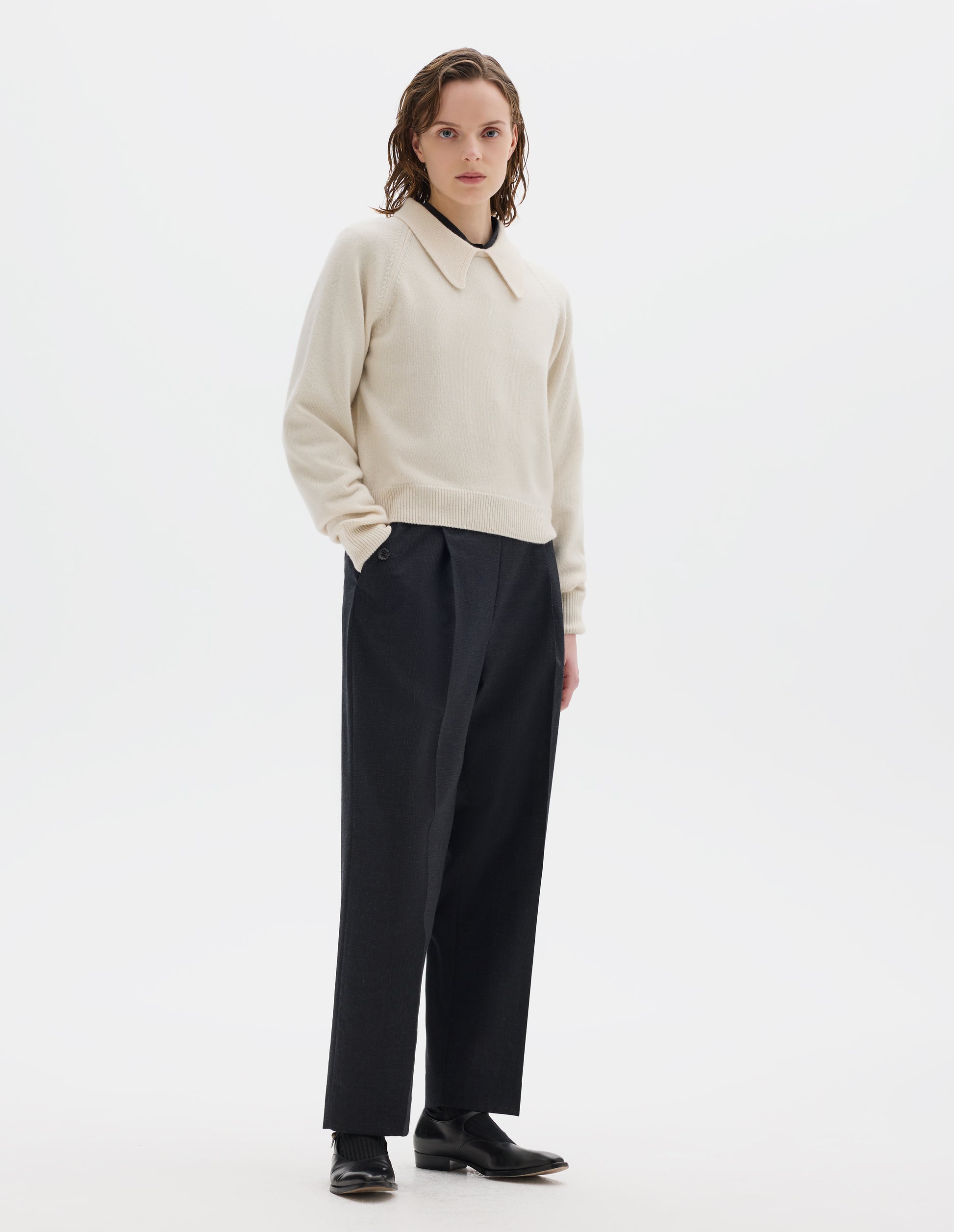 Full body image, front view of model wearing light beige cropped merino jumper with exaggerated collar, raglan sleeves, and with ribbed cuffs and hem. Styled over black shirt, and with dark grey trousers, black socks and black leather shoes.