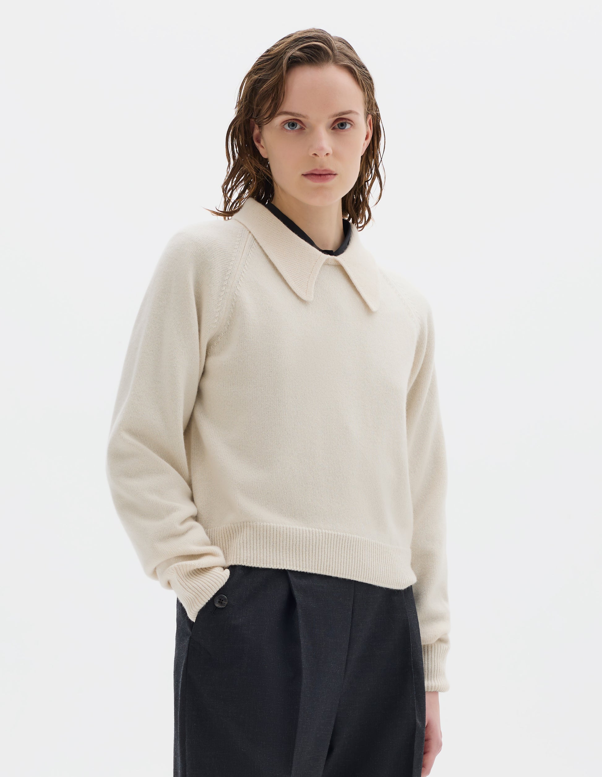 Cropped body image, front view of model wearing light beige cropped merino jumper with exaggerated collar, raglan sleeves, and with ribbed cuffs and hem. Styled over black shirt, and with dark grey trousers.