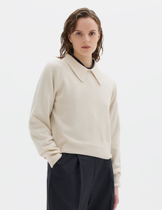 Cropped body image, front view of model wearing light beige cropped merino jumper with exaggerated collar, raglan sleeves, and with ribbed cuffs and hem. Styled over black shirt, and with dark grey trousers.