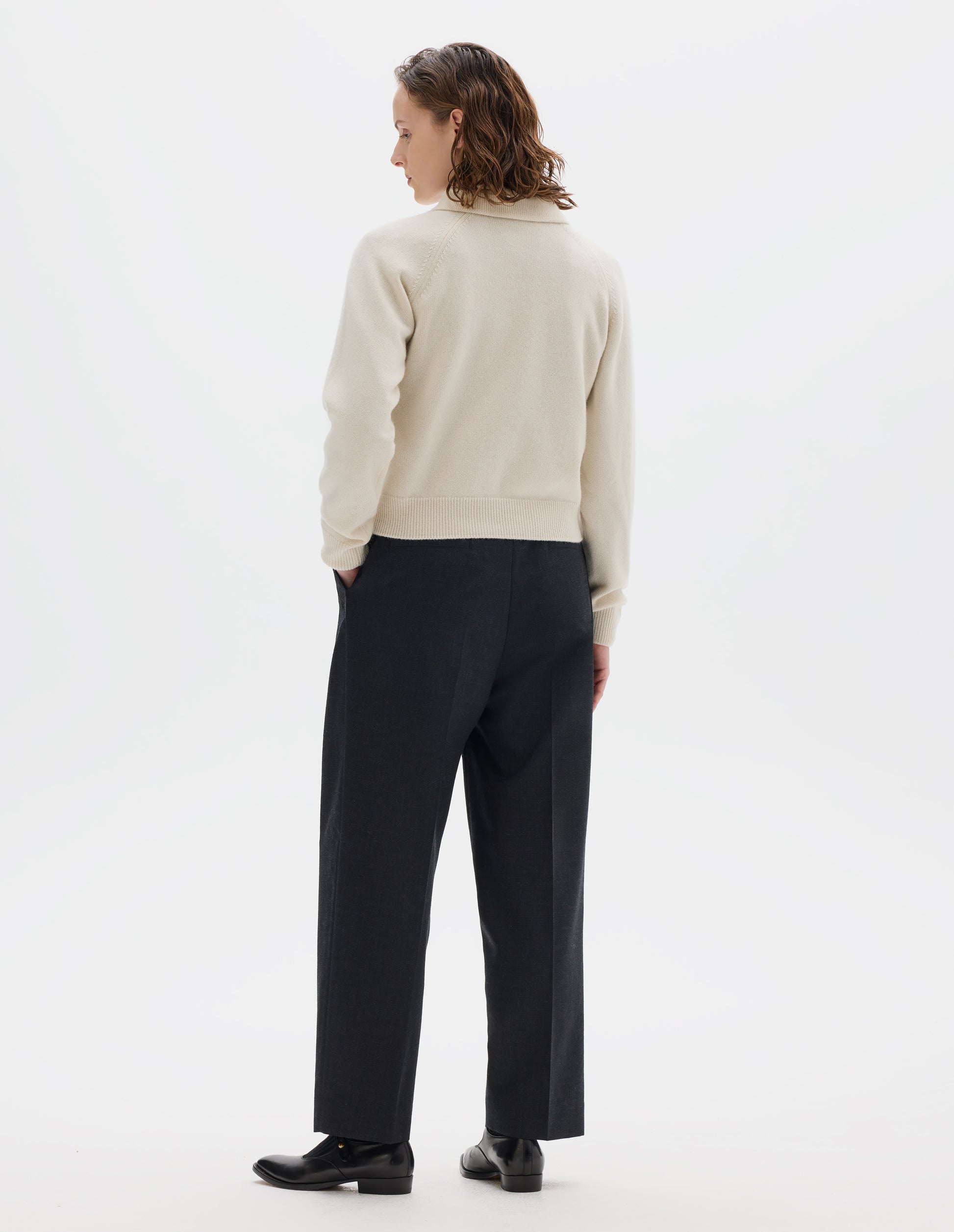 Full body image, back view of model wearing light beige cropped merino jumper with collar, raglan sleeves, and with ribbed cuffs and hem. Styled over black shirt, and with dark grey trousers, black socks and black leather shoes.