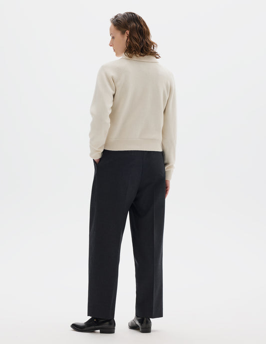 Full body image, back view of model wearing light beige cropped merino jumper with collar, raglan sleeves, and with ribbed cuffs and hem. Styled over black shirt, and with dark grey trousers, black socks and black leather shoes.