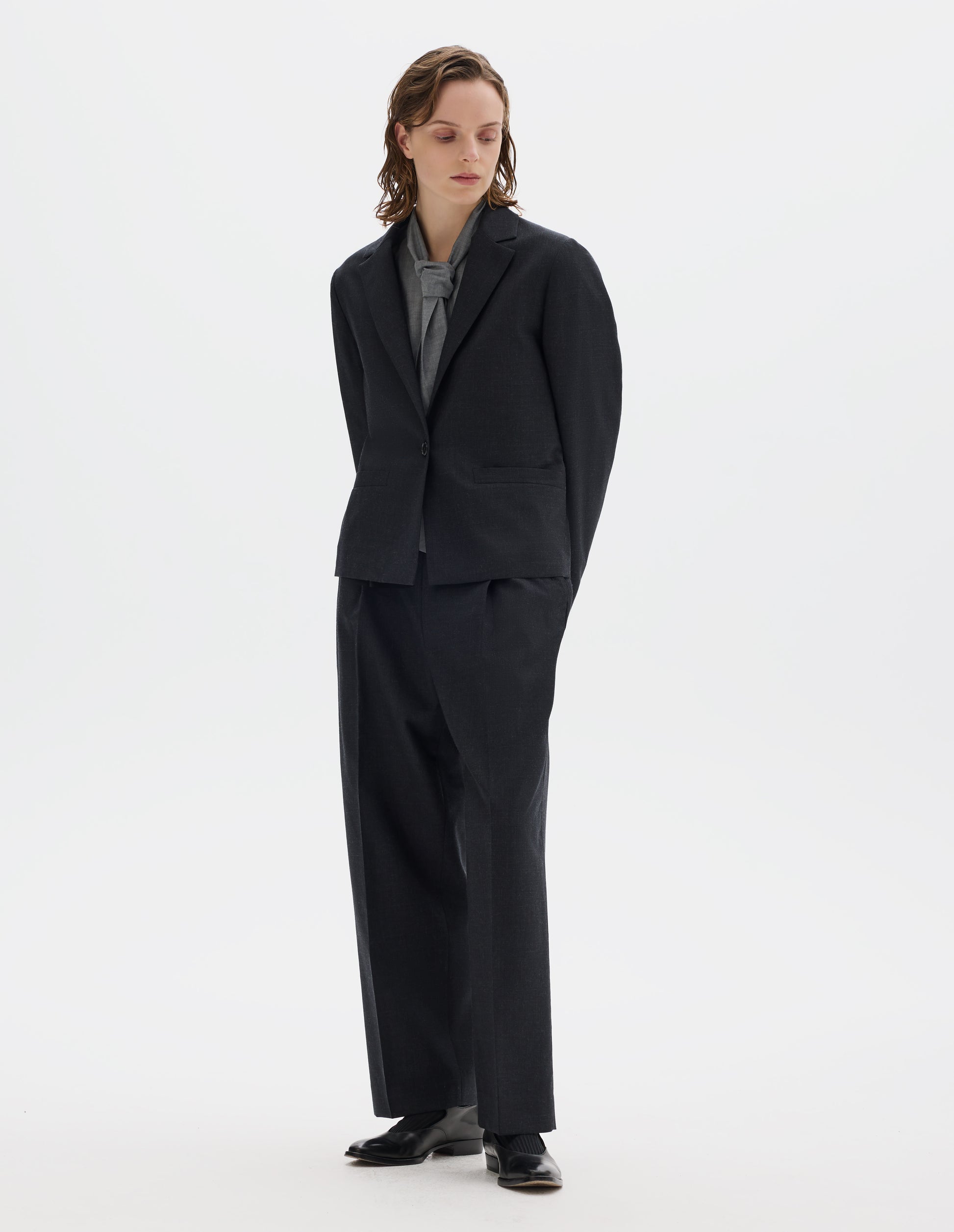 Full body image, front view of model wearing dark grey wool blazer with single horn button fastening, welt pockets and horn buttons on cuffs. Styled over grey shirt, and with matching grey trousers and black leather shoes.
