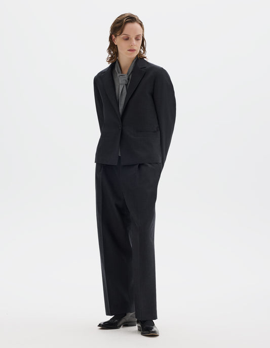Full body image, front view of model wearing dark grey wool blazer with single horn button fastening, welt pockets and horn buttons on cuffs. Styled over grey shirt, and with matching grey trousers and black leather shoes.