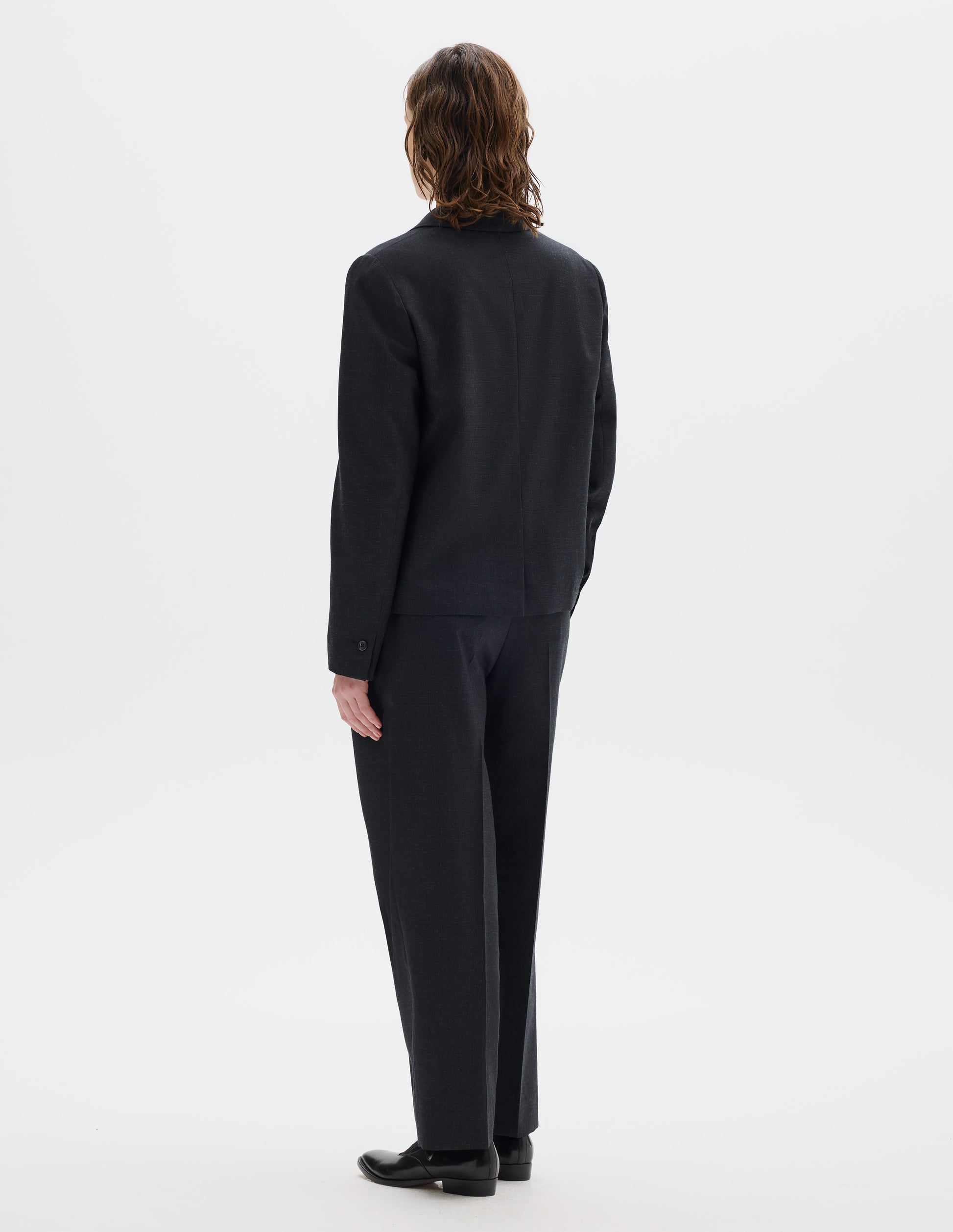 Full body image, back view of model wearing dark grey wool blazer with centre back vent and horn buttons on cuffs. Styled with matching trousers and black leather shoes.