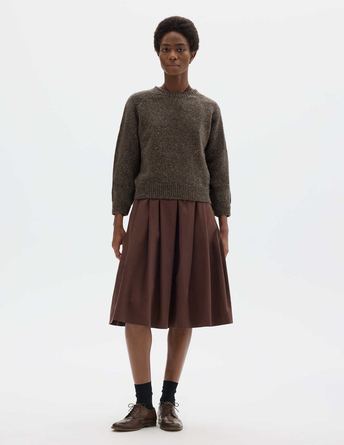 Full body image, front view of model wearing waist length brown crew neck wool jumper with fully fashioned bracelet length raglan sleeves, circular knitted body and wide rib detailing at neck, cuffs and hem. Styled over brown dress, and with black socks and brown leather brogues.