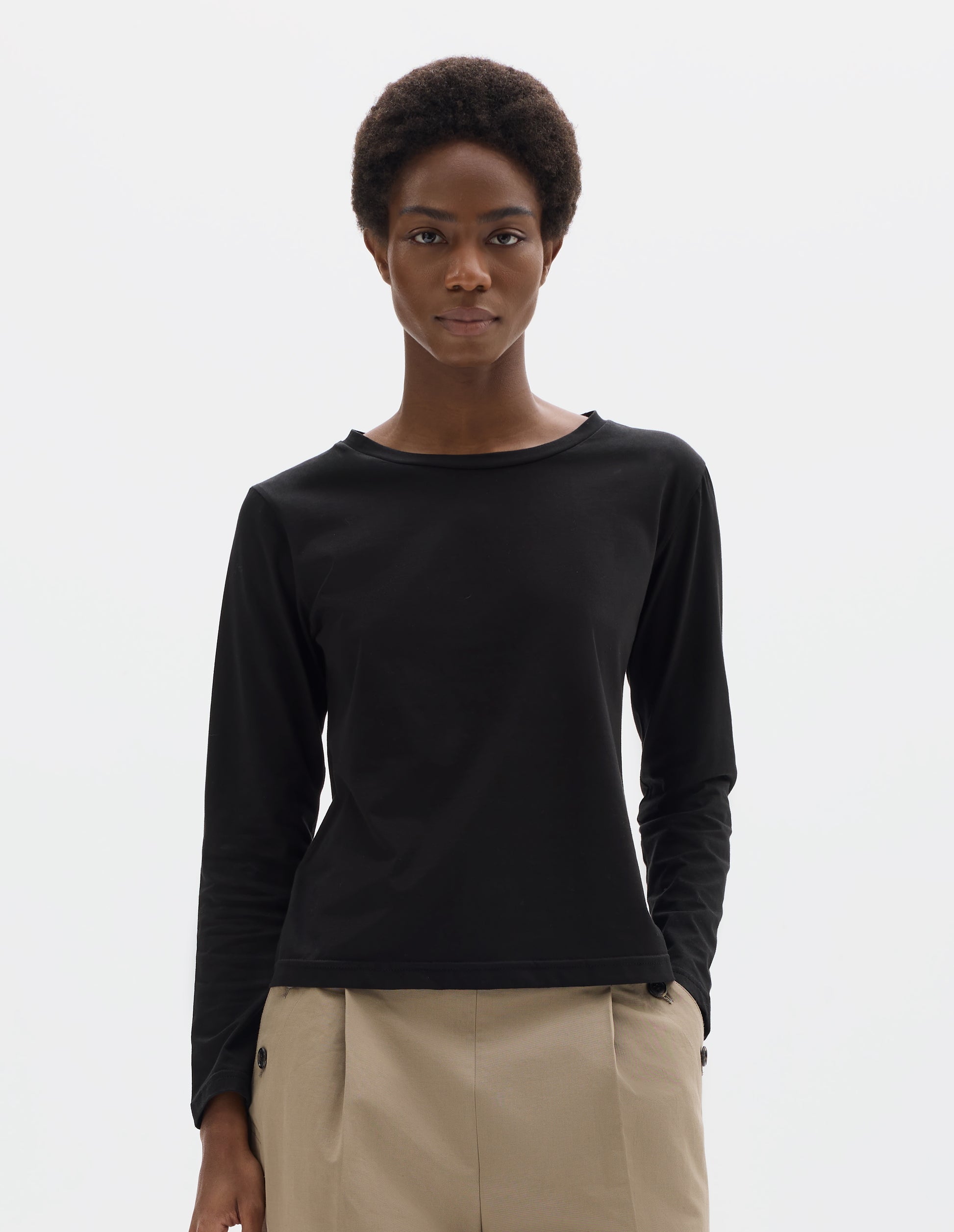 Cropped body image, front view of model wearing black relaxed cotton t-shirt with wide round neck, long sleeves and boxy cut. Styled with beige trousers.