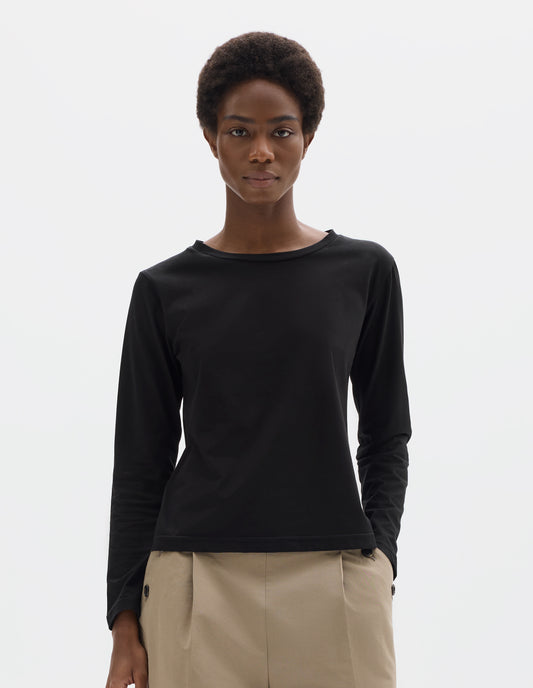 Cropped body image, front view of model wearing black relaxed cotton t-shirt with wide round neck, long sleeves and boxy cut. Styled with beige trousers.