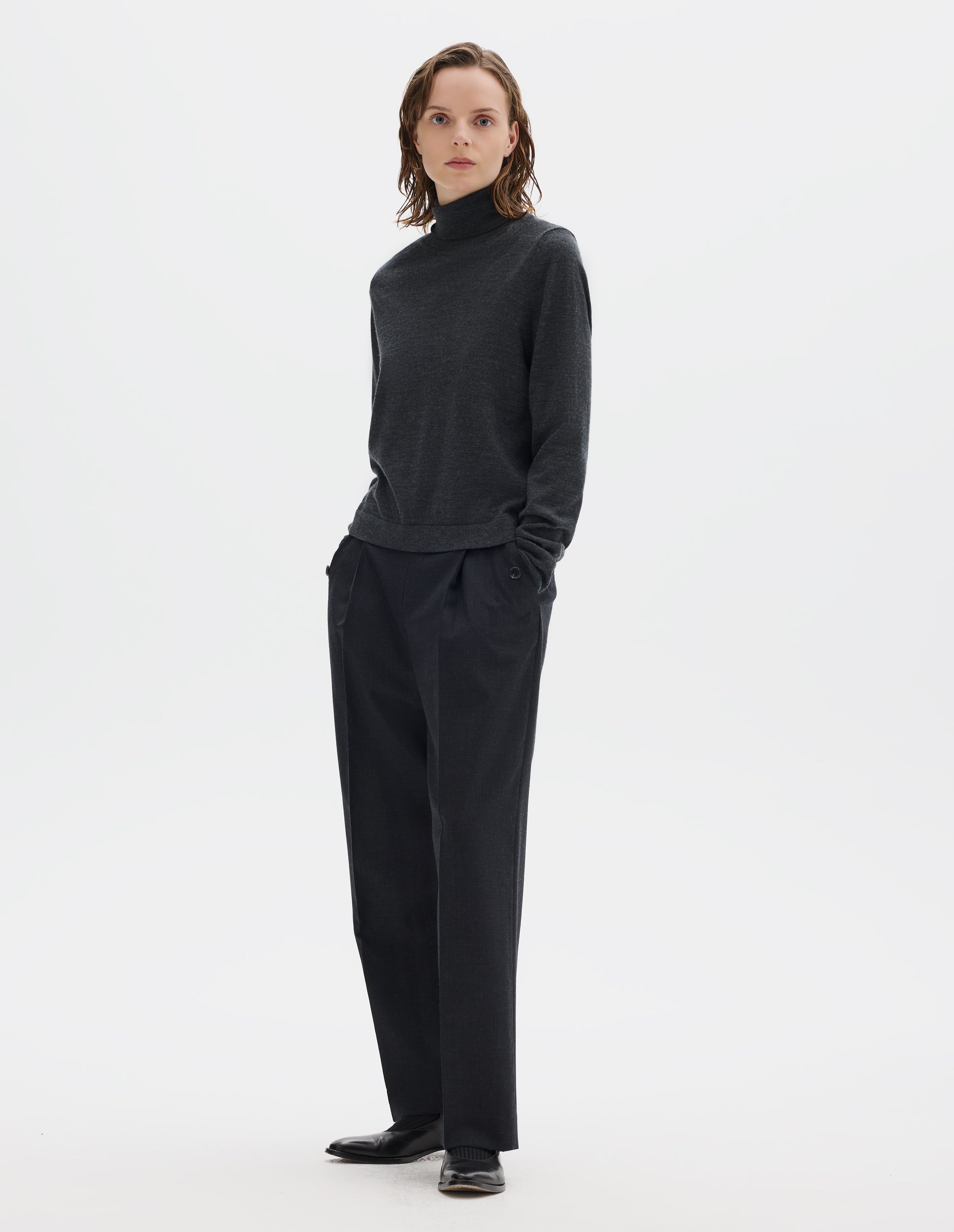 Full body image, front view of model wearing dark grey merino knitted top with fully fashioned roll neck and armhole, and with ribbed neck, sleeves, cuff and hem. Styled with dark grey trousers and black leather shoes.