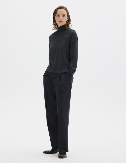 Full body image, front view of model wearing dark grey merino knitted top with fully fashioned roll neck and armhole, and with ribbed neck, sleeves, cuff and hem. Styled with dark grey trousers and black leather shoes.