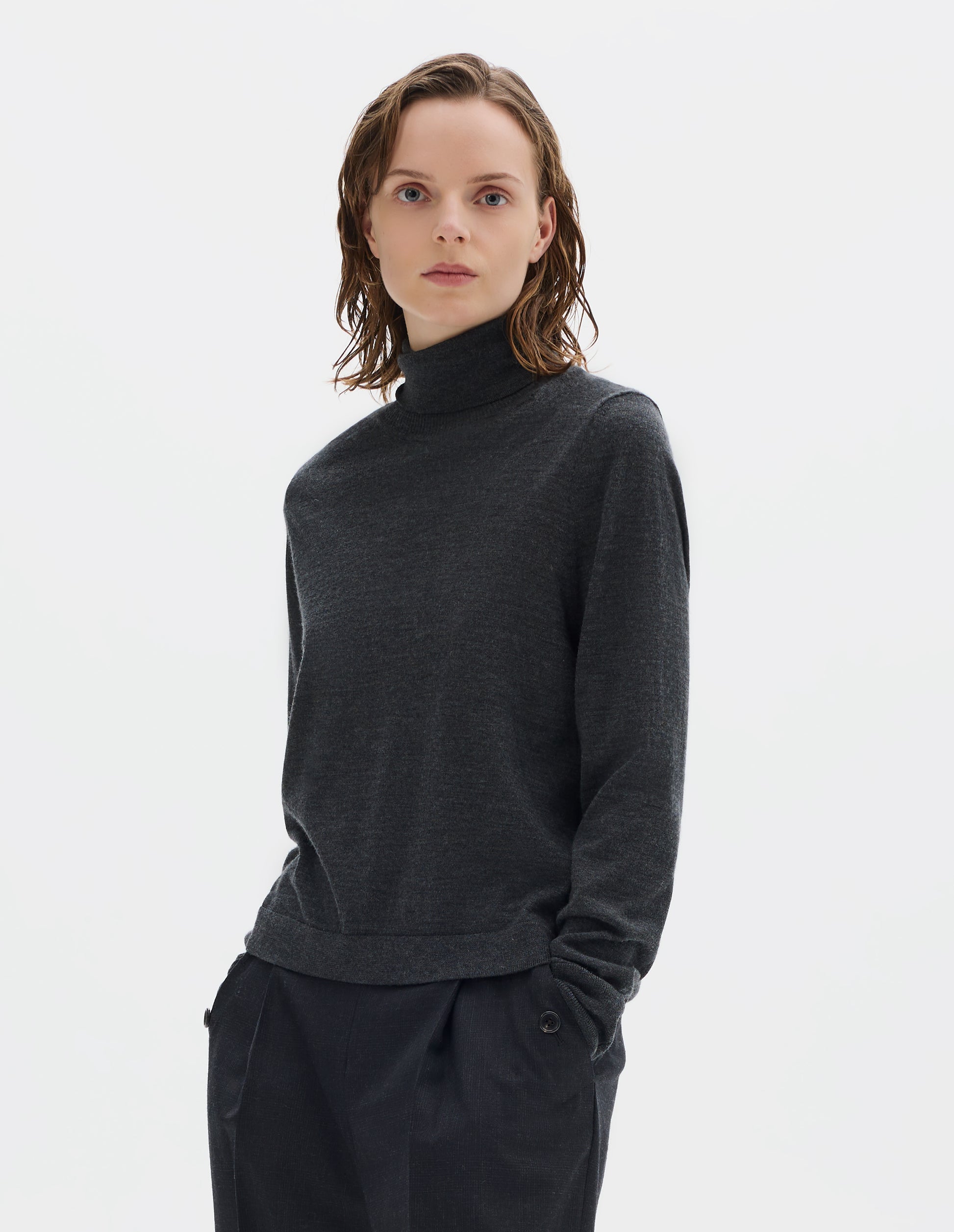 Cropped body image, front view of model wearing dark grey merino knitted top with fully fashioned roll neck and armhole, and with ribbed neck, sleeves, cuff and hem. Styled with dark grey trousers.