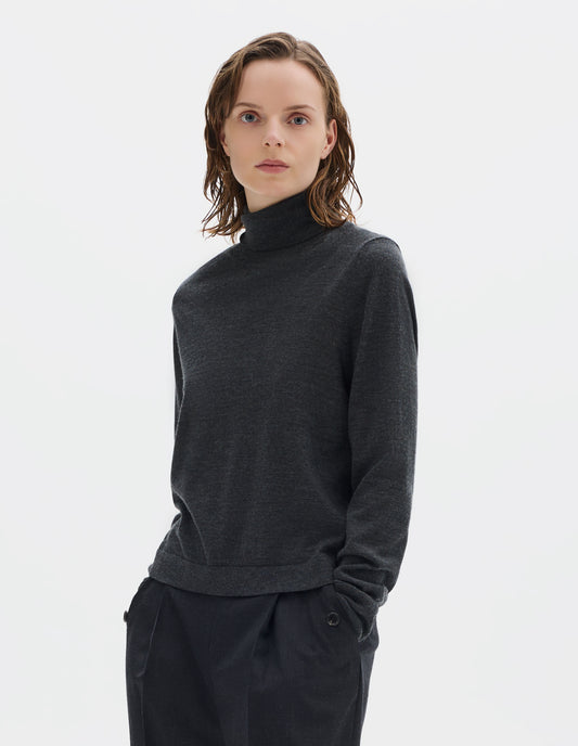 Cropped body image, front view of model wearing dark grey merino knitted top with fully fashioned roll neck and armhole, and with ribbed neck, sleeves, cuff and hem. Styled with dark grey trousers.