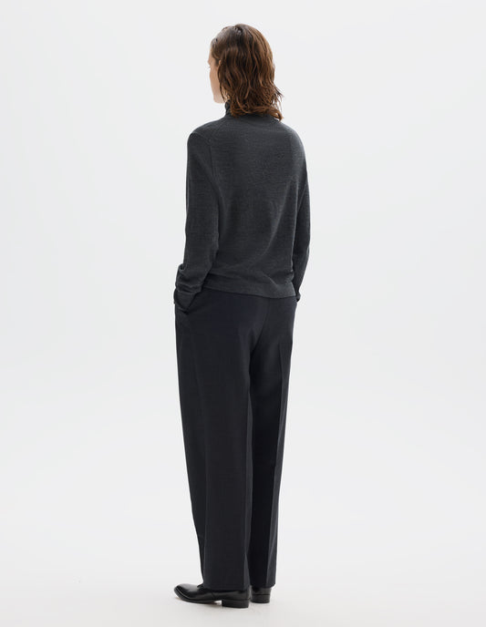 Full body image, back view of model wearing dark grey merino knitted top with fully fashioned roll neck and armhole, and with ribbed neck, sleeves, cuff and hem. Styled with dark grey trousers and black leather shoes.