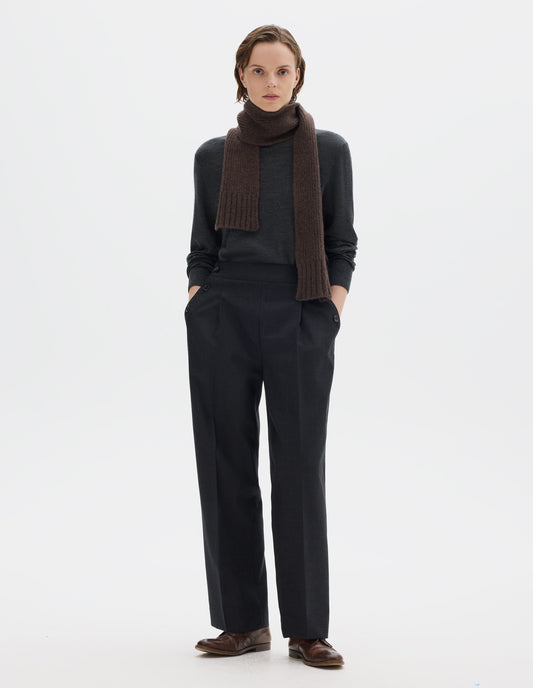 Full body image, front view of model wearing dark grey wool trousers with pleats, horn button side cinches and three horn button closures on both sides, and with stitch-through hem. Styled with brown knitted scarf, dark grey jumper and brown leather brogues.