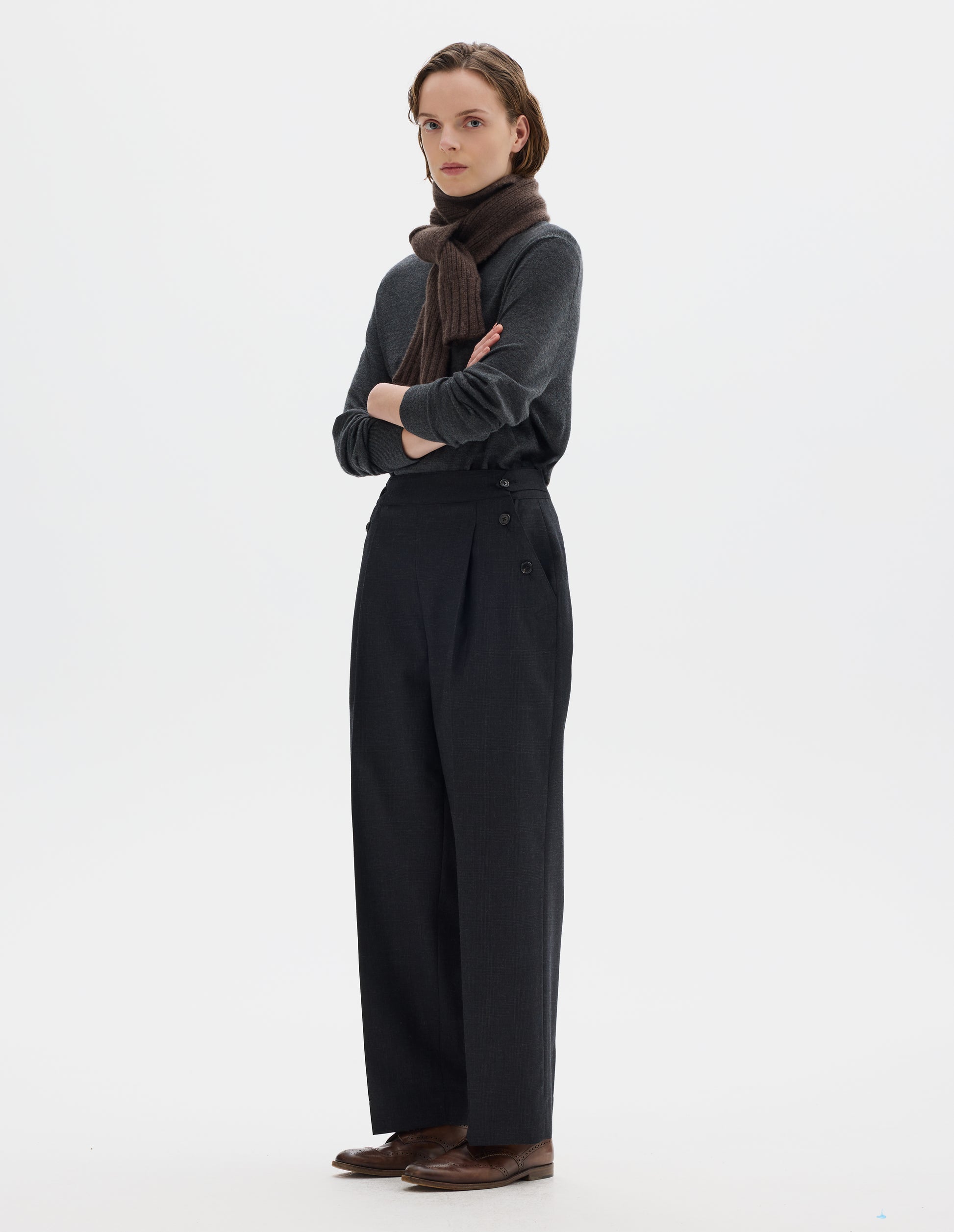 Full body image, side view of model wearing dark grey wool trousers with pleats, horn button side cinches and three horn button closures on both sides, and with stitch-through hem. Styled with brown knitted scarf, dark grey jumper and brown leather brogues.
