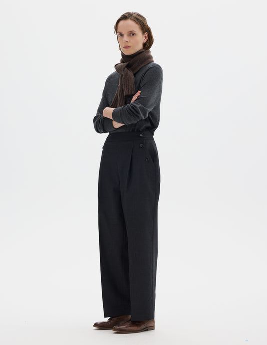 Full body image, side view of model wearing dark grey wool trousers with pleats, horn button side cinches and three horn button closures on both sides, and with stitch-through hem. Styled with brown knitted scarf, dark grey jumper and brown leather brogues.