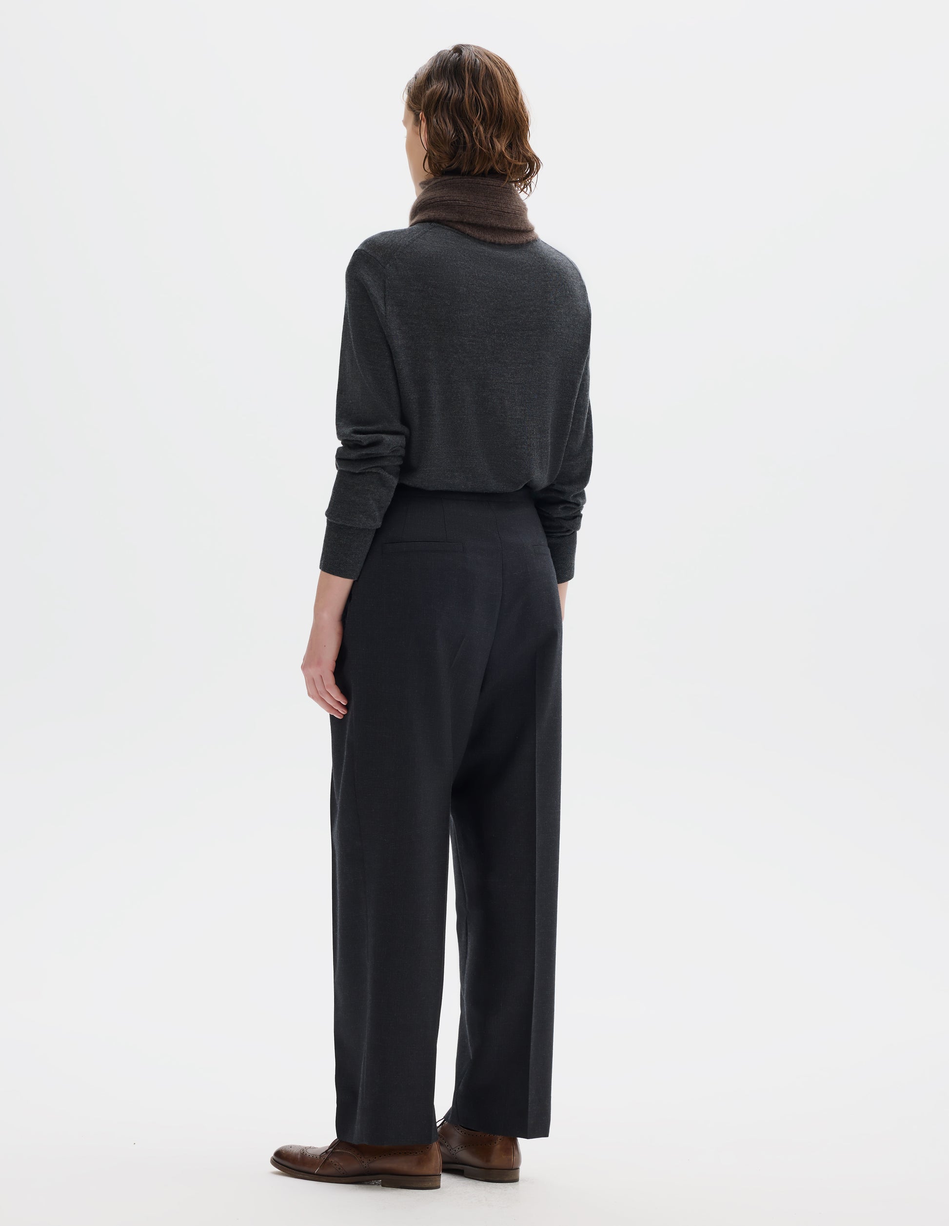 Full body image, back view of model wearing dark grey wool trousers with side cinches and welt back pockets. Styled with brown knitted scarf, dark grey jumper and brown leather brogues.