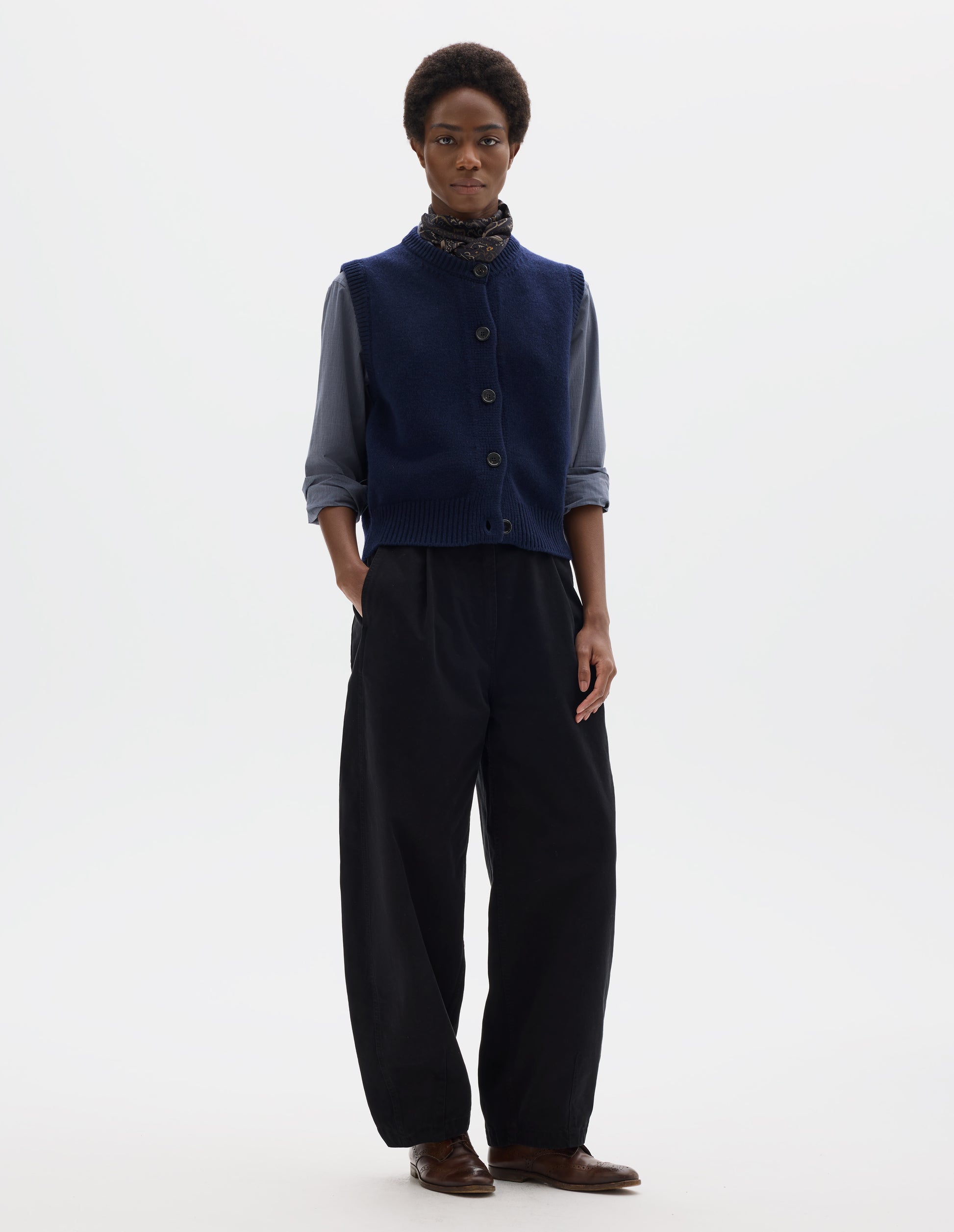 Full body image, front view of model wearing relaxed dark blue cashmere merino knitted gilet with horn buttons on Milano stitch placket, rib detailing at neck, armholes and hem. Styled over blue shirt, and with multicolour silk neck scarf, black trousers and brown leather brogues.
