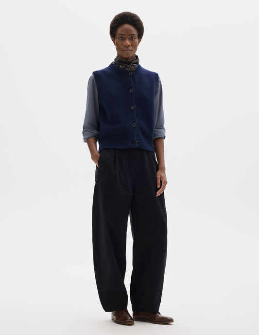 Full body image, front view of model wearing relaxed dark blue cashmere merino knitted gilet with horn buttons on Milano stitch placket, rib detailing at neck, armholes and hem. Styled over blue shirt, and with multicolour silk neck scarf, black trousers and brown leather brogues.