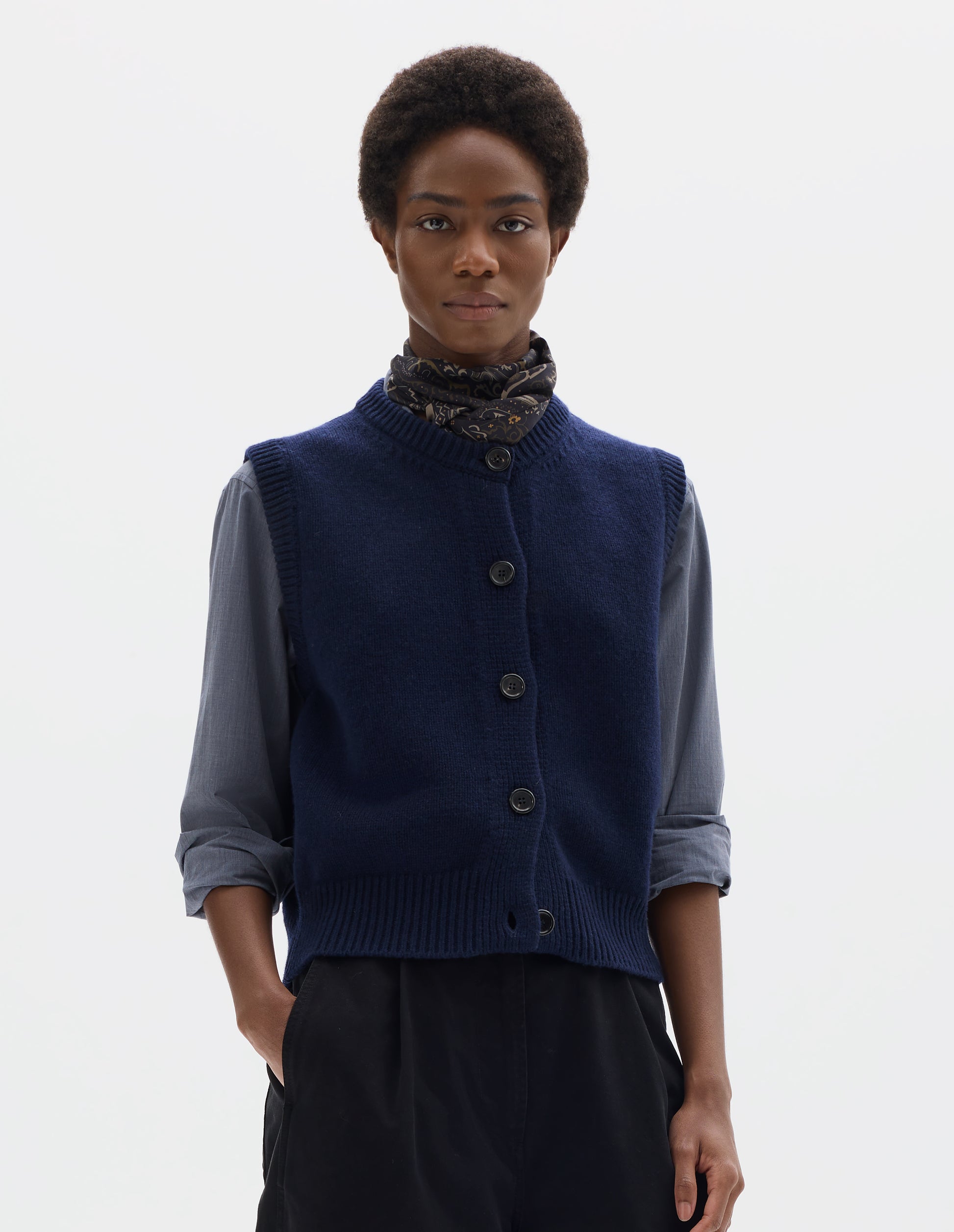 Cropped body image,  front view of model wearing relaxed dark blue cashmere merino knitted gilet with horn buttons on Milano stitch placket, rib detailing at neck, armholes and hem. Styled over blue shirt, and with multicolour silk neck scarf and black trousers.