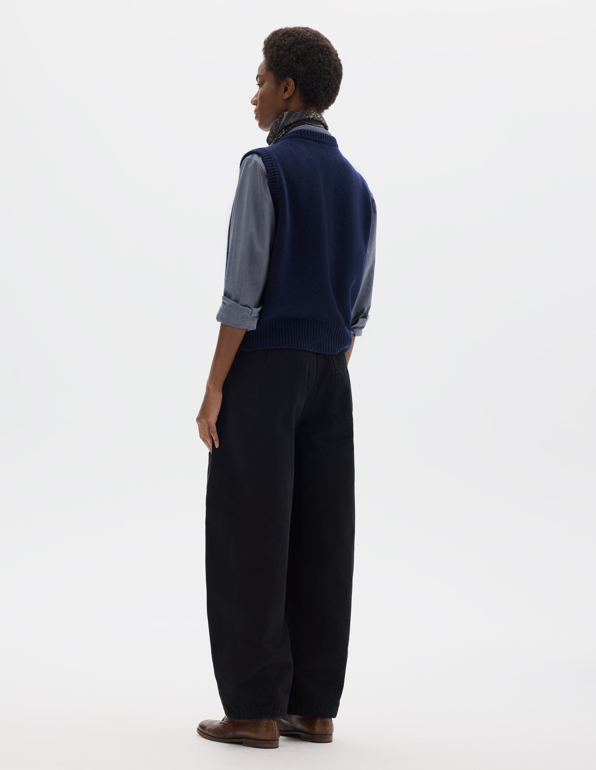 Full body image, back view of model wearing relaxed dark blue cashmere merino knitted gilet with rib detailing at neck, armholes and hem. Styled over blue shirt, and with multicolour silk neck scarf, black trousers and brown leather brogues.