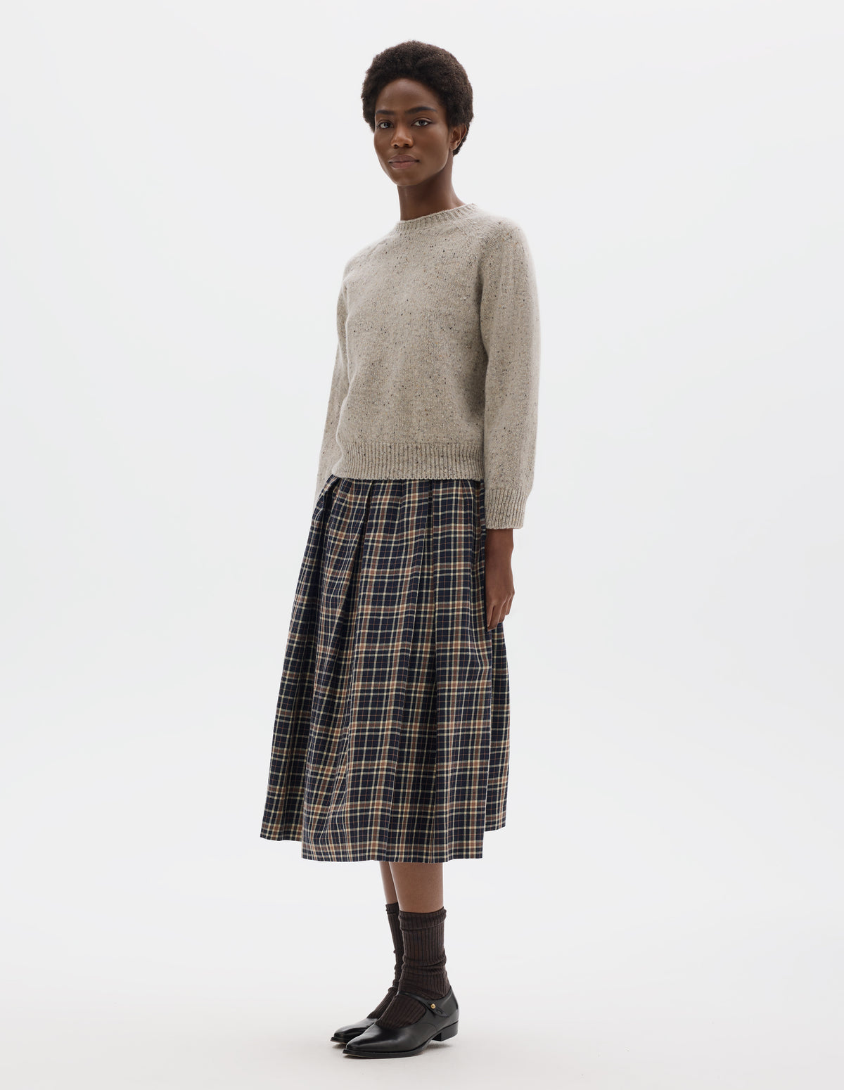 Full body image, front view of model wearing waist length light grey crew neck wool jumper with fully fashioned cropped length raglan sleeves, circular knitted body and wide rib detailing at neck, cuffs and hem. Styled with dark blue and beige checked skirt, brown socks and black leather shoes.