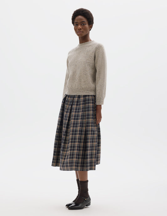 Full body image, front view of model wearing waist length light grey crew neck wool jumper with fully fashioned cropped length raglan sleeves, circular knitted body and wide rib detailing at neck, cuffs and hem. Styled with dark blue and beige checked skirt, brown socks and black leather shoes.