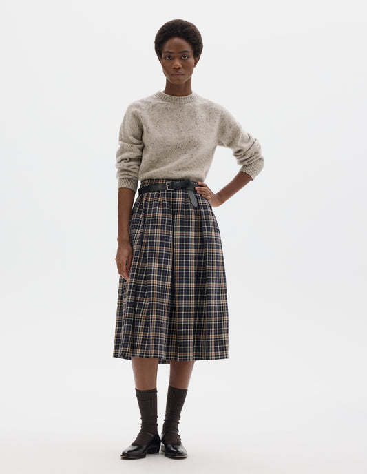 Full body image, front view of model wearing midi length dark blue wool linen skirt with beige and brown checks, narrow waistband, concealed side zip with hook and bar fastening, box pleats with darts, and concealed side seam pockets. Styled with beige jumper, black leather belt, dark brown socks and black leather shoes.