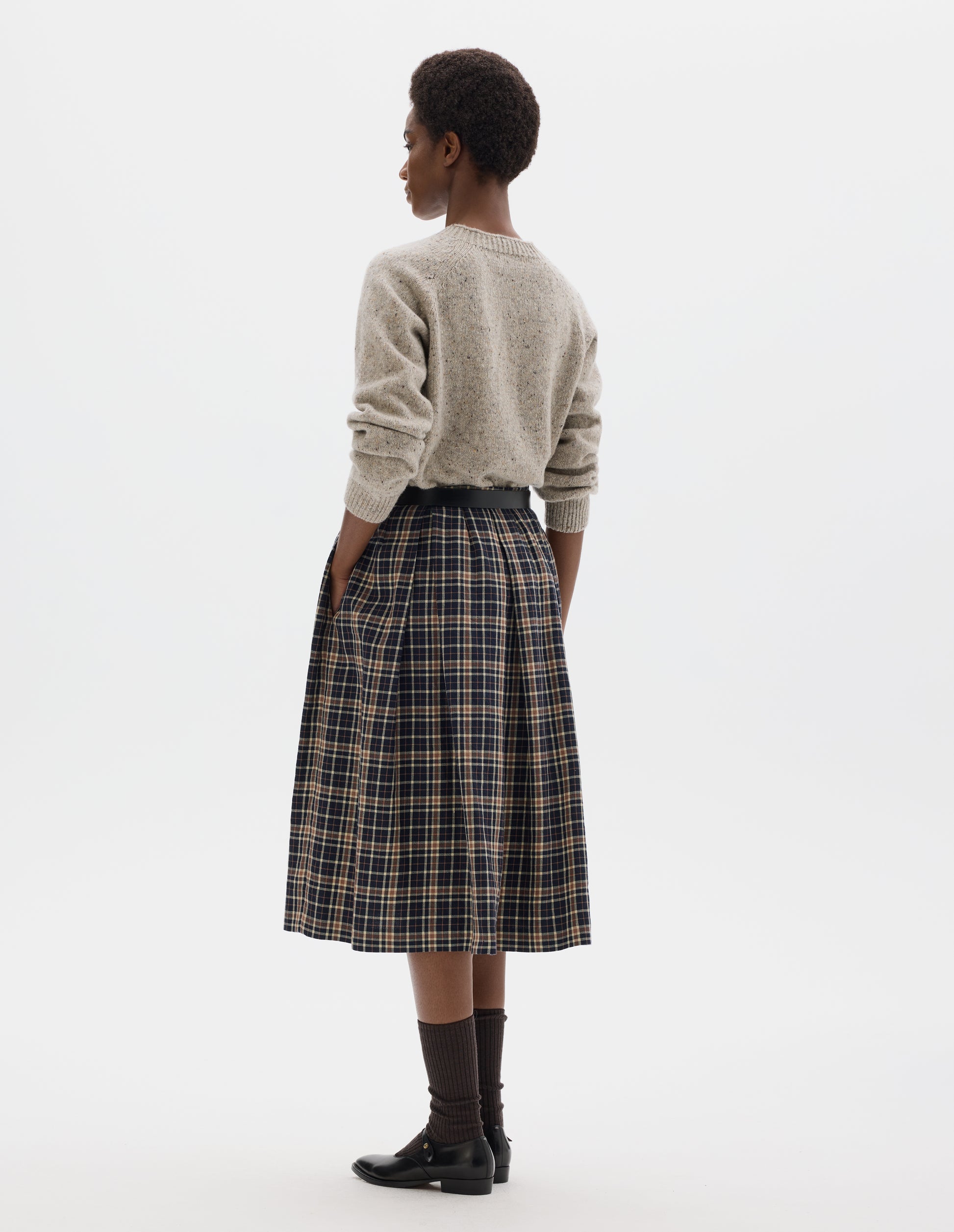 Full body image, back view of model wearing midi length dark blue wool linen skirt with beige and brown checks, narrow waistband, concealed side zip with hook and bar fastening, box pleats with darts, and concealed side seam pockets. Styled with beige jumper, black leather belt, dark brown socks and black leather shoes.
