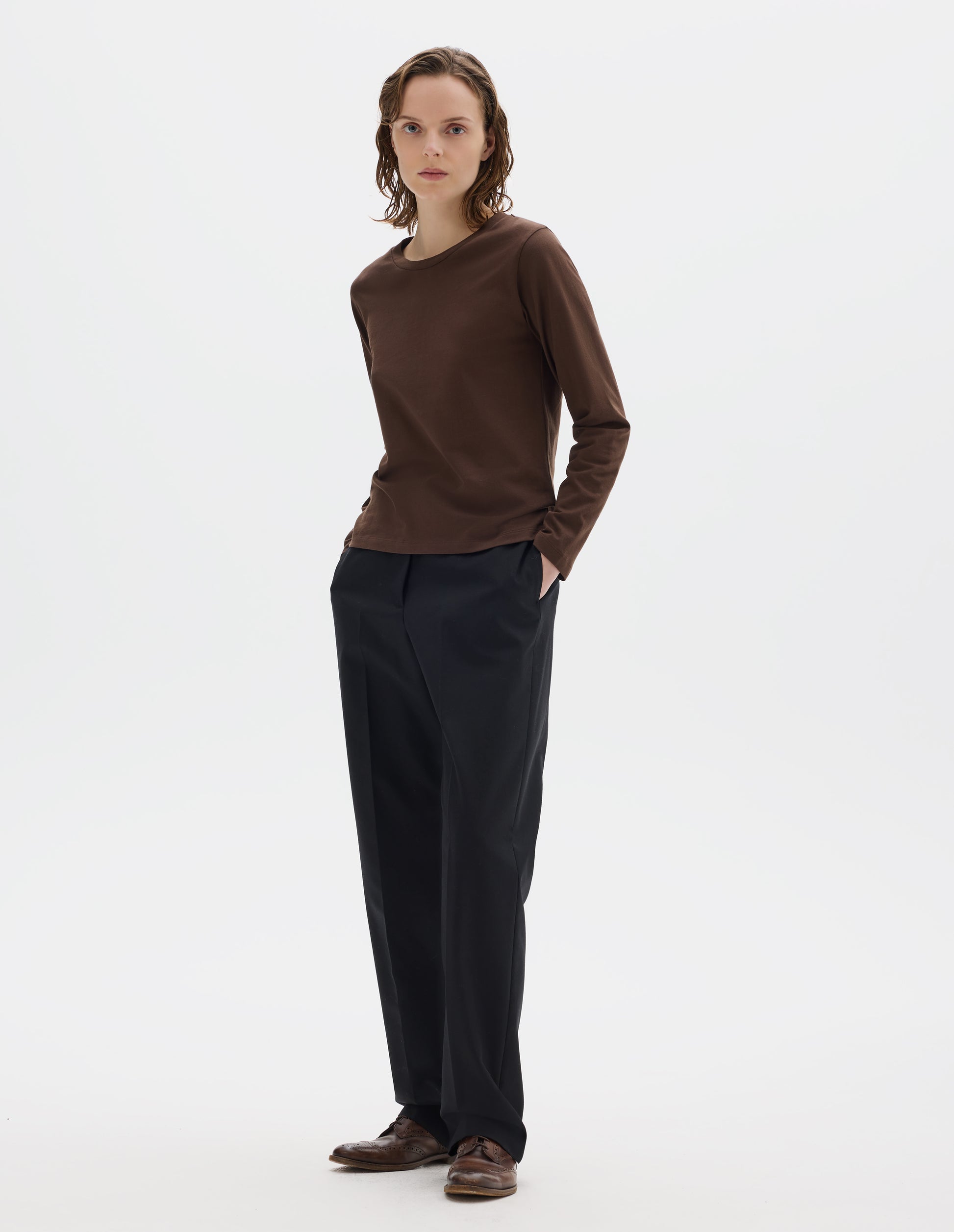 Full body image, front view of model wearing relaxed dark brown jersey t-shirt with boxy cut, wide neck and long sleeves. Styled with black trousers and brown leather brogues.