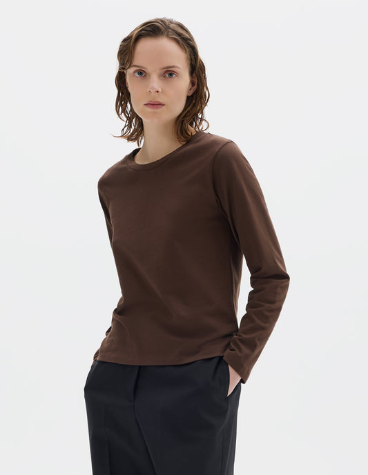 Cropped body image, front view of model wearing relaxed dark brown jersey t-shirt with boxy cut, wide neck and long sleeves. Styled with black trousers.