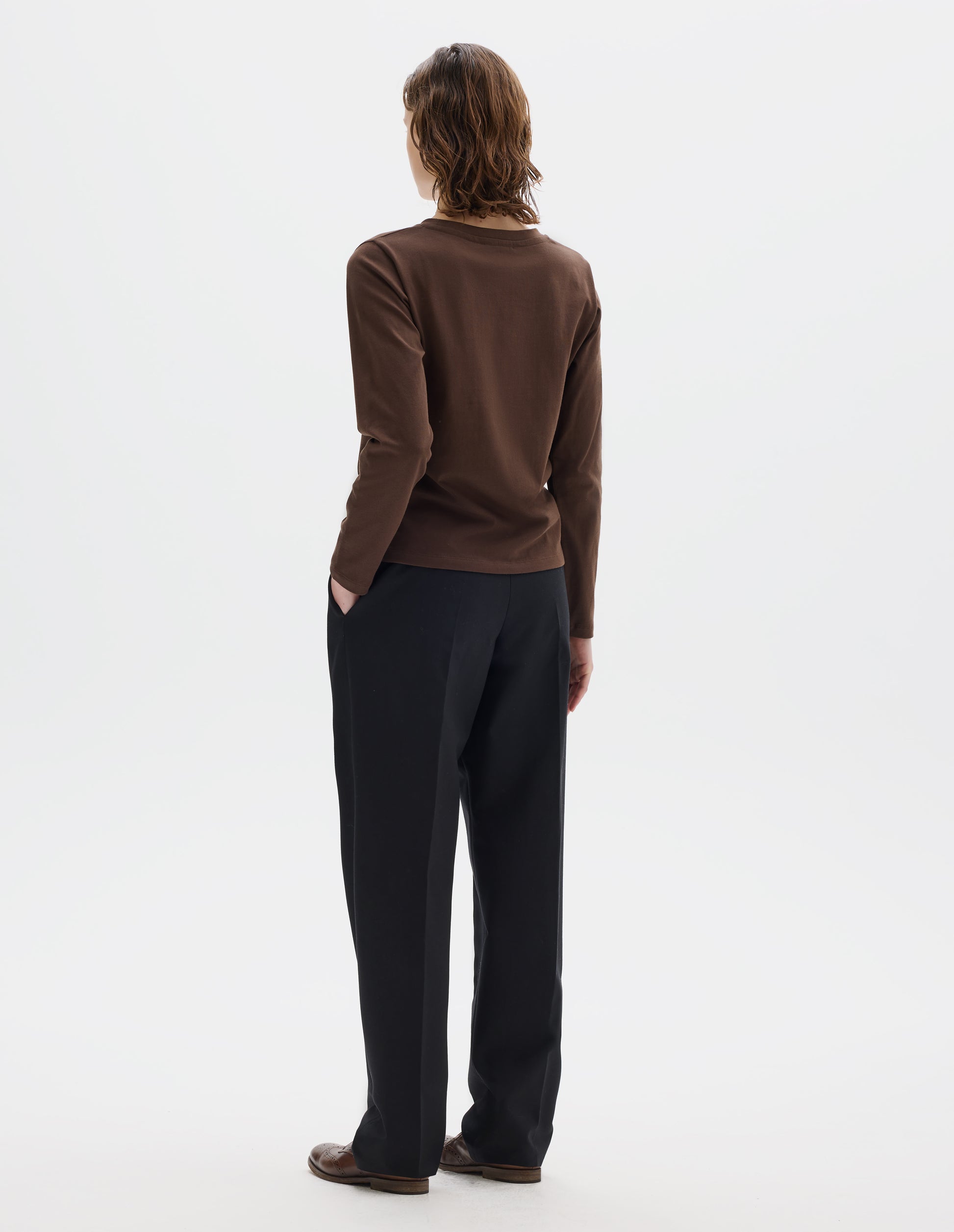 Full body image, back view of model wearing relaxed dark brown jersey t-shirt with boxy cut, wide neck and long sleeves. Styled with black trousers and brown leather brogues.