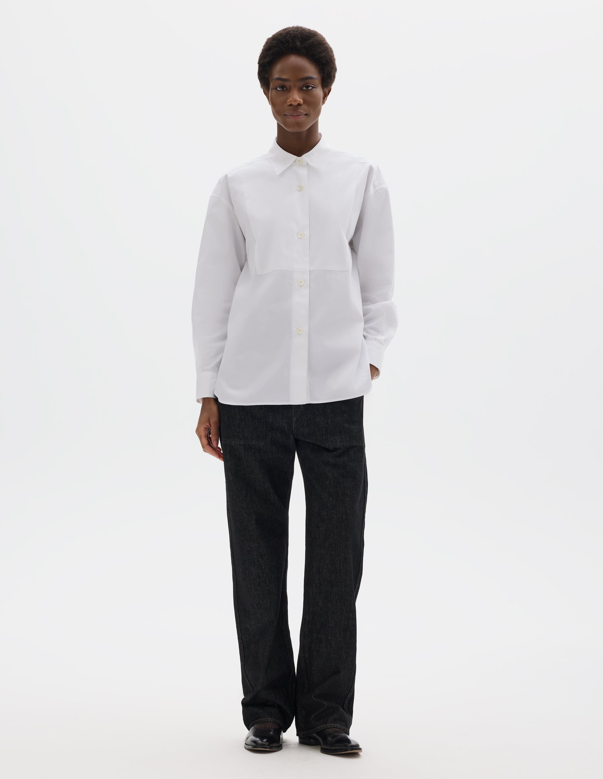 Full body image, front view of model wearing white oversized cotton shirt with pointed stand collar, mother of pearl buttons on placket, double-layer bib front and curved hem. Styled with black jeans and black leather shoes.