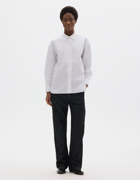 Full body image, front view of model wearing white oversized cotton shirt with pointed stand collar, mother of pearl buttons on placket, double-layer bib front and curved hem. Styled with black jeans and black leather shoes.